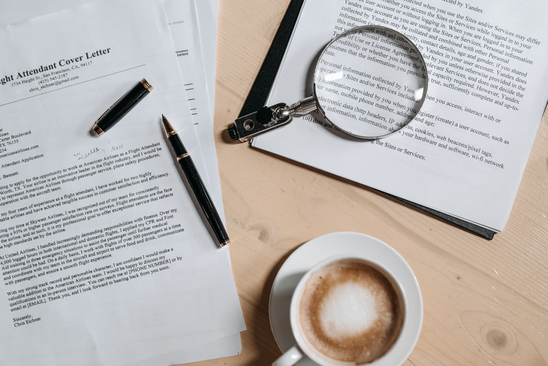 A top view of coffee and job application documents with a magnifying glass on a wooden desk.